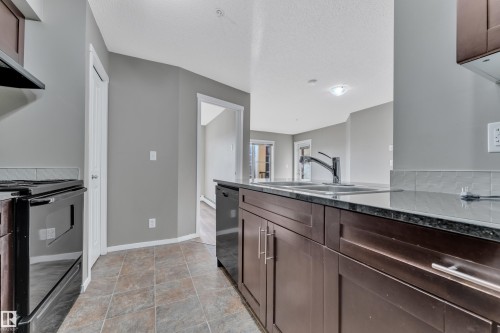 Kitchen with dark wood finish cabinetry, stainless steel electric range, dark stone counters, dishwashing machine, and a textured ceiling - 216 5804 Mullen Place, Edmonton, AB - Indoor Photo Showing Kitchen With Double Sink