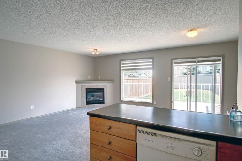 Kitchen with dishwasher, a textured ceiling, carpet, dark countertops, and a fireplace - 8129 7 Avenue, Edmonton, AB - Indoor With Fireplace