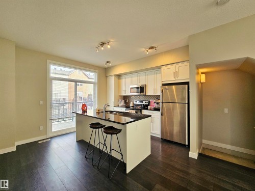 Kitchen with stainless steel appliances, an island with sink, a breakfast bar, dark wood-type flooring, and white cabinets - 129 2560 Pegasus Boulevard, Edmonton, AB - Indoor Photo Showing Kitchen