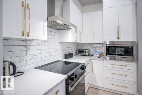 Kitchen featuring black electric range oven, wall chimney range hood, white cabinetry, decorative backsplash, and stainless steel microwave - 101 7471 May Common, Edmonton, AB - Indoor Photo Showing Kitchen With Upgraded Kitchen