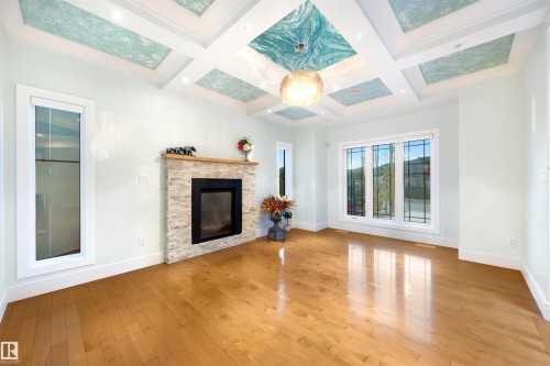 Unfurnished living room featuring light wood-style flooring, a stone fireplace, coffered ceiling, and crown molding - 4811 Woolsey Lane, Edmonton, AB - Indoor Photo Showing Other Room With Fireplace