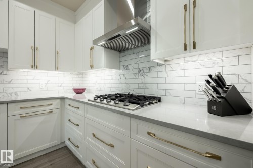 Kitchen featuring white cabinets, wall chimney range hood, backsplash, light stone countertops, and stainless steel gas cooktop - 7471 May Common, Edmonton, AB - Indoor Photo Showing Kitchen With Upgraded Kitchen