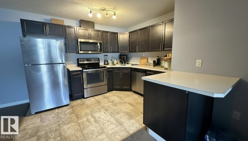 Kitchen featuring stainless steel appliances, light countertops, a peninsula, a textured ceiling, and dark wood finish cabinets - 133 301 Clareview Station Drive, Edmonton, AB - Indoor Photo Showing Kitchen