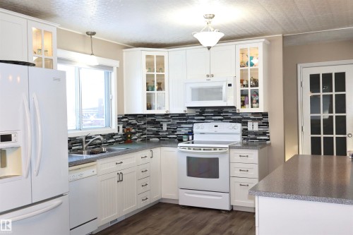 Kitchen with white appliances, white cabinetry, dark countertops, dark wood finished floors, and glass fronted cabinets - 5220 - 56 Street, Barrhead, AB - Indoor Photo Showing Kitchen With Double Sink