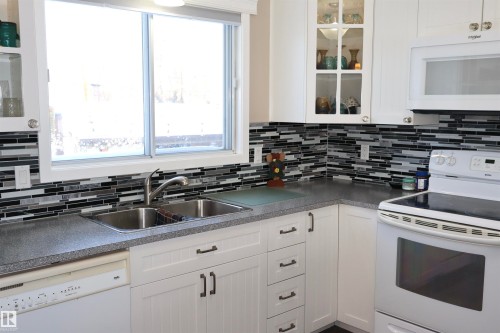 Kitchen with white appliances, dark countertops, glass fronted cabinets, and white cabinetry - 5220 - 56 Street, Barrhead, AB - Indoor Photo Showing Kitchen With Double Sink