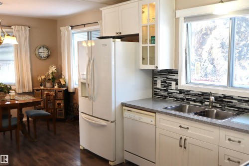 Kitchen with decorative light fixtures, white cabinetry, white appliances, dark countertops, and glass insert cabinets - 5220 - 56 Street, Barrhead, AB - Indoor Photo Showing Kitchen With Double Sink