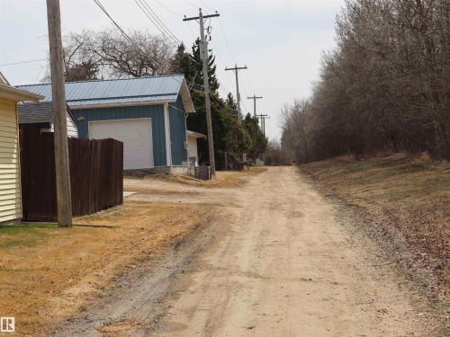 Detached blue outbuilding with a metal roof and white garage door, featuring a brown privacy fence and mature trees - 5220 - 56 Street, Barrhead, AB - Outdoor