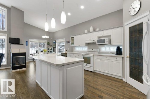 Kitchen featuring white cabinets, white appliances, pendant lighting, dark wood-style floors, and a multi sided fireplace - 8 Osborne Close, St. Albert, AB - Indoor Photo Showing Kitchen With Fireplace