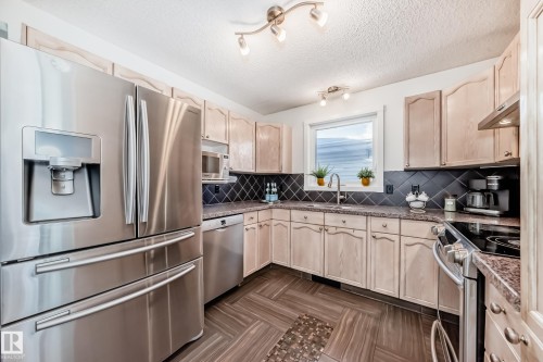 Kitchen featuring stainless steel appliances, light wood finish cabinets, a textured ceiling, dark stone countertops, and tasteful backsplash - 7011 190B Street, Edmonton, AB - Indoor Photo Showing Kitchen With Double Sink