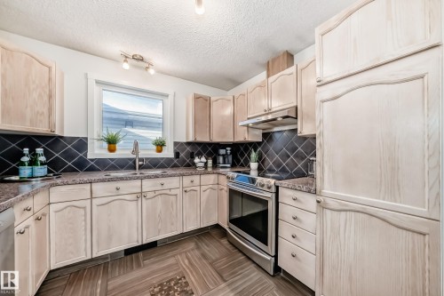Kitchen with stainless steel appliances, light wood finish cabinetry, a textured ceiling, dark stone counters, and tasteful backsplash - 7011 190B Street, Edmonton, AB - Indoor Photo Showing Kitchen With Double Sink