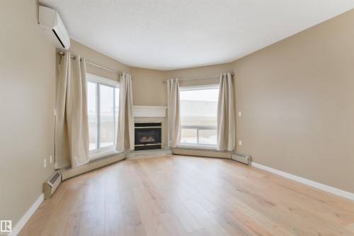 Unfurnished living room featuring a glass covered fireplace, a baseboard heating unit, a baseboard radiator, light wood finished floors, and healthy amount of natural light - 232 15499 Castle Downs Road, Edmonton, AB - Indoor Photo Showing Other Room With Fireplace
