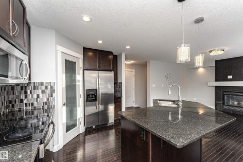 Kitchen featuring dark wood finish cabinets, dark stone countertops, stainless steel appliances, hanging light fixtures, and dark wood-type flooring - 6963 19A Avenue, Edmonton, AB - Indoor Photo Showing Kitchen With Upgraded Kitchen