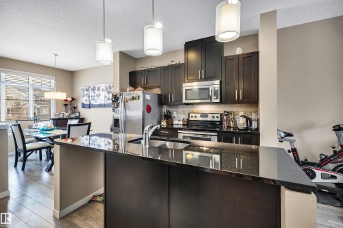 Kitchen featuring hanging light fixtures, dark stone counters, stainless steel appliances, dark wood finish cabinetry, and light wood finished floors - 8 320 Secord Boulevard, Edmonton, AB - Indoor Photo Showing Kitchen With Double Sink With Upgraded Kitchen