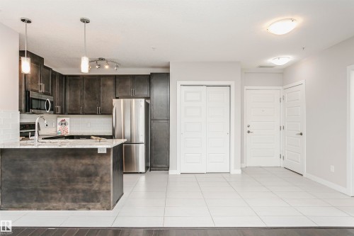 Kitchen with dark wood finish cabinets, stainless steel appliances, light stone counters, a peninsula, and light tile patterned floors - 332 5151 Windermere Boulevard, Edmonton, AB - Indoor Photo Showing Kitchen