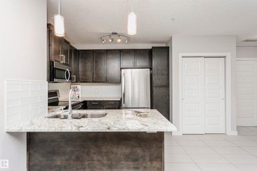 Kitchen featuring stainless steel appliances, light stone countertops, dark wood finish cabinetry, a peninsula, and pendant lighting - 332 5151 Windermere Boulevard, Edmonton, AB - Indoor Photo Showing Kitchen