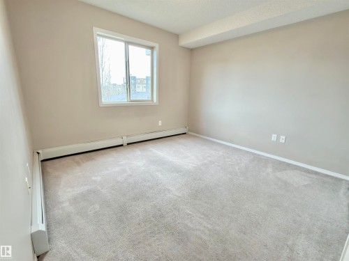 This room features light-colored walls and carpeting, a window providing natural light, and baseboard heating - 205 11816 22 Avenue, Edmonton, AB - Indoor Photo Showing Other Room