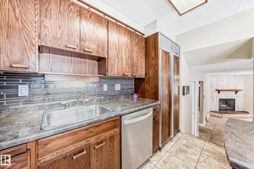 Kitchen featuring dishwasher, a textured ceiling, a stone fireplace, wood finish cabinets, and light tile patterned floors - 38 Lancaster Crescent, St. Albert, AB - Indoor Photo Showing Kitchen