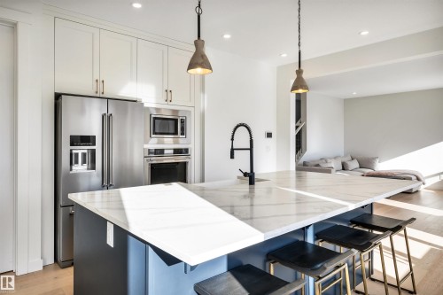 Dual tone kitchen featuring light wood-style floors, stainless steel appliances, a kitchen breakfast bar, hanging light fixtures, and light stone counters - 11 Promenade Point(E), Beaumont, AB - Indoor Photo Showing Kitchen With Upgraded Kitchen
