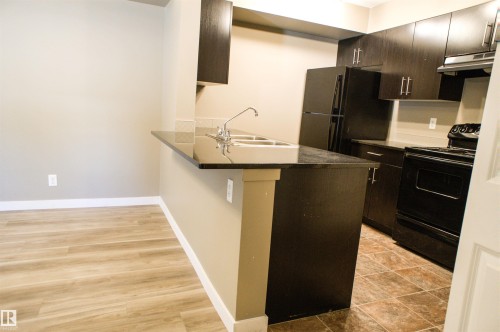 Kitchen with black appliances, a peninsula, a kitchen breakfast bar, dark wood finish cabinetry, and light wood-style floors - 104 5804 Place Nw, Edmonton, AB - Indoor Photo Showing Kitchen With Double Sink With Upgraded Kitchen