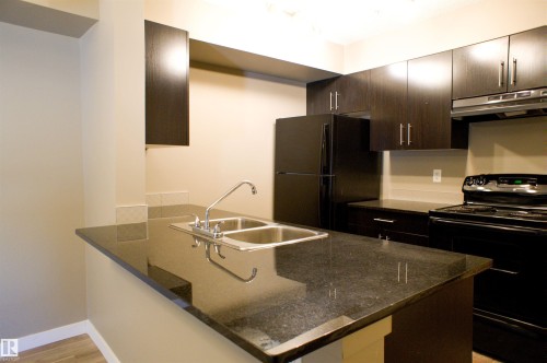 Kitchen with black appliances, dark wood finish cabinets, a peninsula, dark stone countertops, and light wood-style flooring - 104 5804 Place Nw, Edmonton, AB - Indoor Photo Showing Kitchen With Double Sink