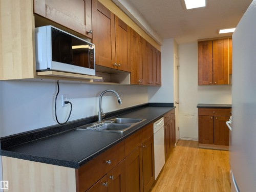 Kitchen with wood finish cabinets, light wood-type flooring, dark countertops, and white appliances - 109 7220 144 Avenue, Edmonton, AB - Indoor Photo Showing Kitchen With Double Sink