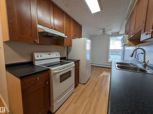 Kitchen with white appliances, dark countertops, light wood finished floors, a textured ceiling, and wood finish cabinets - 109 7220 144 Avenue, Edmonton, AB - Indoor Photo Showing Kitchen With Double Sink