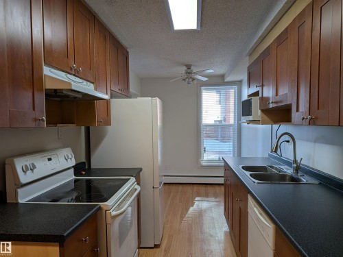 Kitchen featuring white appliances, wood finish cabinets, dark countertops, light wood-type flooring, and a textured ceiling - 109 7220 144 Avenue, Edmonton, AB - Indoor Photo Showing Kitchen With Double Sink