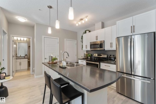 Kitchen with stainless steel appliances, a breakfast bar area, white cabinets, a center island with sink, and a textured ceiling - 103 320 Ambleside Link Link Sw, Edmonton, AB - Indoor Photo Showing Kitchen With Stainless Steel Kitchen With Double Sink With Upgraded Kitchen