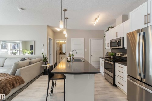 Kitchen featuring stainless steel appliances, white cabinetry, decorative light fixtures, a kitchen breakfast bar, and open floor plan - 103 320 Ambleside Link Link Sw, Edmonton, AB - Indoor Photo Showing Kitchen With Stainless Steel Kitchen With Double Sink With Upgraded Kitchen