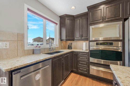 Kitchen with dark wood finish cabinetry, stainless steel appliances, light stone countertops, a warming drawer, and light wood-style flooring - 281 Southwick Way, Leduc, AB - Indoor Photo Showing Kitchen With Double Sink