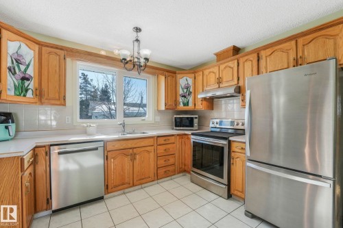 Kitchen with stainless steel appliances, light countertops, a chandelier, wood finish cabinets, and a textured ceiling - 760 Knottwood Road S, Edmonton, AB - Indoor Photo Showing Kitchen With Double Sink