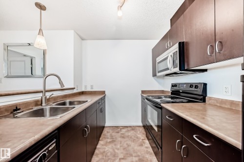 Kitchen featuring black appliances, hanging light fixtures, dark wood finish cabinetry, a textured ceiling, and light countertops - 306 1188 Hyndman Road, Edmonton, AB - Indoor Photo Showing Kitchen With Double Sink