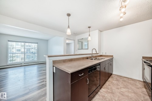 Kitchen featuring a peninsula, dishwasher, dark wood finish cabinetry, electric stove, and light countertops - 306 1188 Hyndman Road, Edmonton, AB - Indoor Photo Showing Kitchen With Double Sink