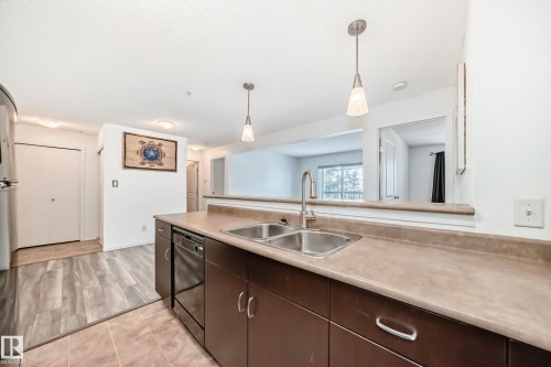 Kitchen featuring dark wood finish cabinets, decorative light fixtures, dishwasher, and light countertops - 306 1188 Hyndman Road, Edmonton, AB - Indoor Photo Showing Kitchen With Double Sink