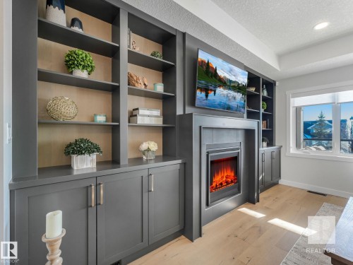 Living room featuring built in shelves, light wood-style floors, a textured ceiling, and a warm lit fireplace - 209 55101 Ste. Anne Trail, Rural Lac Ste. Anne County, AB - Indoor With Fireplace