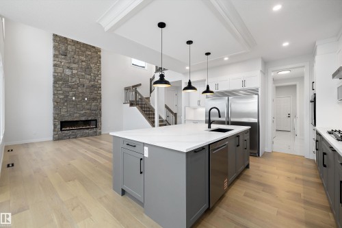 Two tone kitchen featuring dual tone cabinetry, an island with sink, a fireplace, light stone countertops, and light wood finished floors - 718 Elderberry Close Nw, Edmonton, AB - Indoor Photo Showing Kitchen With Upgraded Kitchen