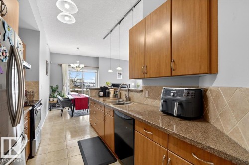 Kitchen featuring wood finish cabinetry, light tile patterned flooring, stainless steel appliances, dark stone counters, and a textured ceiling - 105 9707 105 Street, Edmonton, AB - Indoor Photo Showing Kitchen With Double Sink