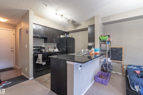 Kitchen featuring black appliances, light carpet, dark cabinets, a textured ceiling, and a breakfast bar - 206 3207 James Mowatt Trail, Edmonton, AB - Indoor Photo Showing Kitchen