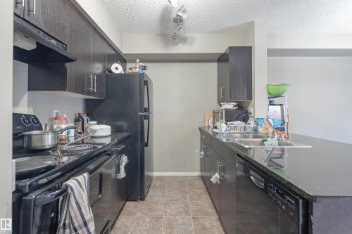 Kitchen featuring black appliances, a textured ceiling, dark stone counters, a peninsula, and rail lighting - 206 3207 James Mowatt Trail, Edmonton, AB - Indoor Photo Showing Kitchen With Double Sink