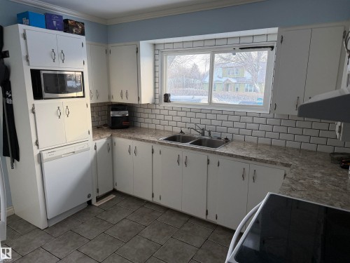 Kitchen featuring decorative backsplash, white cabinetry, white dishwasher, stainless steel microwave, and crown molding - 4506 53 Avenue, Barrhead, AB - Indoor Photo Showing Kitchen With Double Sink
