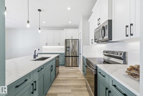 Dual tone kitchen featuring stainless steel appliances, decorative light fixtures, light wood finished floors, tasteful backsplash, and light stone counters - 28 Harley Way, Spruce Grove, AB - Indoor Photo Showing Kitchen With Double Sink With Upgraded Kitchen