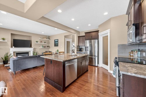 Kitchen with open floor plan, dark wood finish cabinetry, light stone countertops, stainless steel appliances, and a kitchen island with sink - 21 Dittrich, Fort Saskatchewan, AB - Indoor Photo Showing Kitchen With Fireplace With Double Sink With Upgraded Kitchen