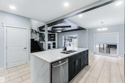 Kitchen featuring dishwasher, light stone countertops, dark cabinets, a center island with sink, and beam ceiling - 