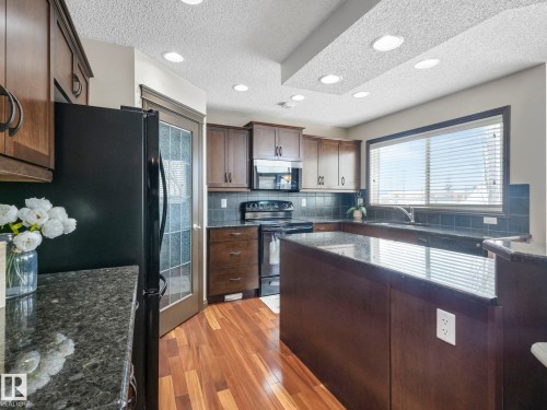 Kitchen with dark wood finish cabinetry, black appliances, light wood-type flooring, dark stone countertops, and backsplash - 960 Summerside Link, Edmonton, AB - Indoor Photo Showing Kitchen