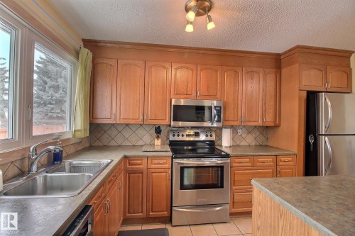 Kitchen featuring stainless steel appliances, light tile patterned floors, wood finish cabinetry, a textured ceiling, and tasteful backsplash - 4520 20A Avenue, Edmonton, AB - Indoor Photo Showing Kitchen With Double Sink