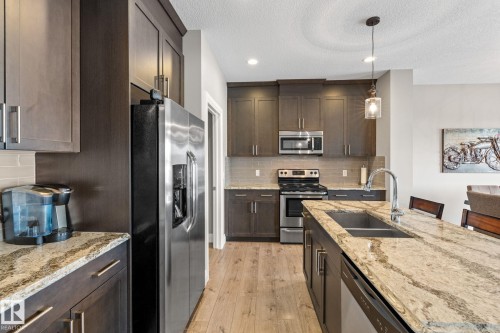 Kitchen featuring light stone counters, dark wood finish cabinetry, stainless steel appliances, light wood-type flooring, and decorative backsplash - 1043 South Creek Wynd, Stony Plain, AB - Indoor Photo Showing Kitchen With Double Sink With Upgraded Kitchen