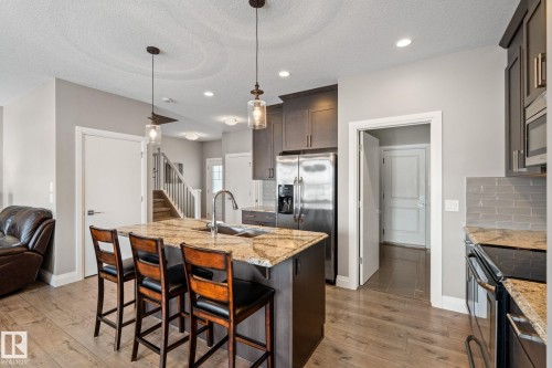 Kitchen featuring backsplash, light stone countertops, a center island with sink, stainless steel appliances, and dark wood finish cabinetry - 1043 South Creek Wynd, Stony Plain, AB - Indoor Photo Showing Kitchen With Upgraded Kitchen