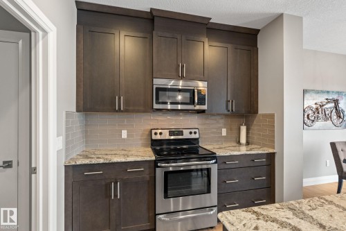 Kitchen with stainless steel appliances, dark wood finish cabinetry, light stone countertops, decorative backsplash, and a textured ceiling - 1043 South Creek Wynd, Stony Plain, AB - Indoor Photo Showing Kitchen With Upgraded Kitchen