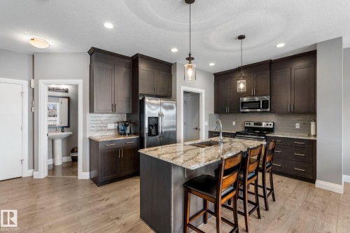 Kitchen featuring dark wood finish cabinets, light stone countertops, stainless steel appliances, an island with sink, and a breakfast bar area - 1043 South Creek Wynd, Stony Plain, AB - Indoor Photo Showing Kitchen With Upgraded Kitchen