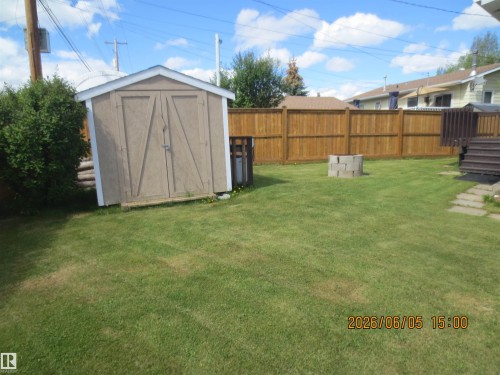 Expansive grassy yard featuring a wood-panel storage shed, wood privacy fencing, a concrete block fire pit, and stone paver stepping stones - 1102 55 Street, Edson, AB 